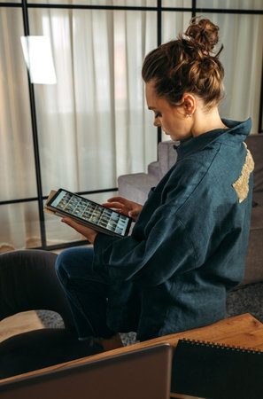 A young woman in a dark blue suit sits on the back of a sofa with a tablet in her hands. Browse online stores, social networks. back view. vertical frame.の写真素材