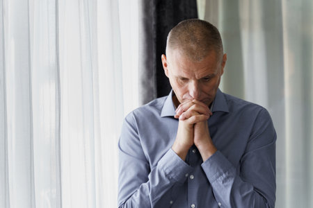 Portrait of a man who stands at the window clenched his hands in front of him, concentrated, prays or thinks.の写真素材