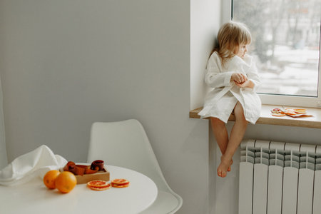 A girl in a white coat is sad sitting on the windowsill, eating fruit marshmallow.の写真素材