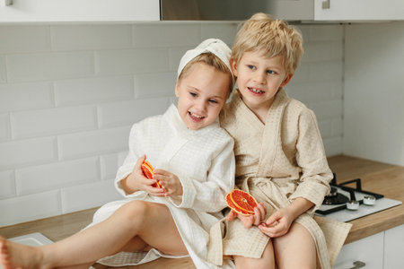 A girl and a boy in bathrobes sit in the kitchen and close their eyes with candied oranges.の写真素材