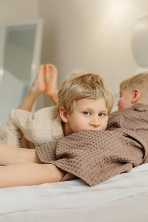 The boys lie on the bed in bathrobes after a shower. The elder put his head on the younger, looks into the camera.の写真素材