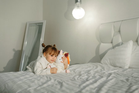 A girl in a bathrobe plays on the bed with a knitted unicorn.の写真素材