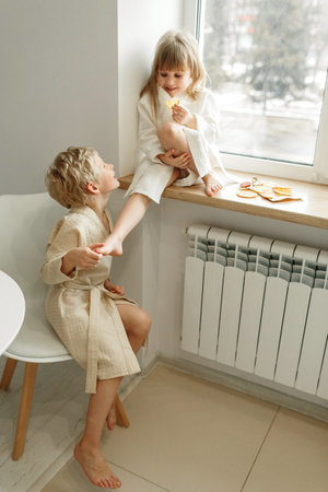 Portrait of a fair-haired smiling boy in a bathrobe who sits on a chair and leans on the girl's leg.の写真素材