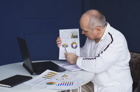 A man makes an online video call chat, sitting at home and talking into a laptop webcam, showing a document.の写真素材