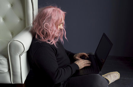 A man in a pink wig sits on the floor near a white chair and works at a computer. side viewの写真素材