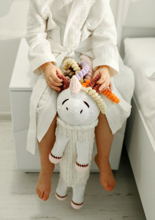 A girl in a bathrobe plays with a knitted unicorn.の写真素材