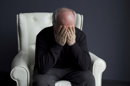 A man in a black turtleneck is sitting in a white chair, rubbing his face with his hands, holding his head. depression, headache. medical concept.の写真素材