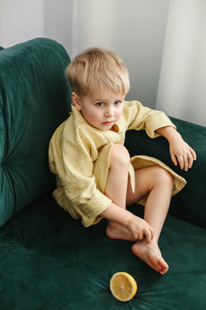 A boy in a yellow bathrobe sits on a green sofa, half an orange lies next to him.の写真素材