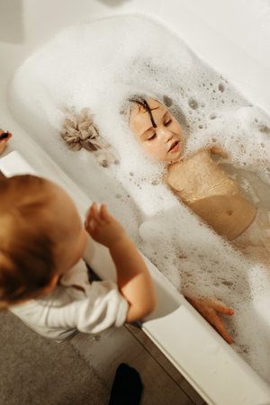 A happy girl swims in a bathtub full of foam with her little sister standing next to her. Only the...