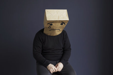 A sad man in a black turtleneck with a bag on his head, with a drawn crying emoticon, sits on a chair. Emotions and gestures.の写真素材