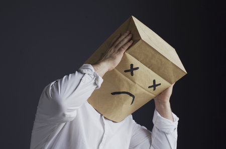A man in a white shirt with a bag on his head, with a sad emoticon drawn, stands holding his head with his hands.の写真素材