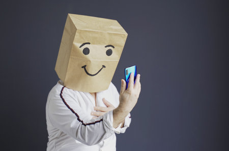 A man in a white shirt with a paper bag on his head, with a smiley face drawn, writes a message and takes a selfie on a smartphone.の写真素材
