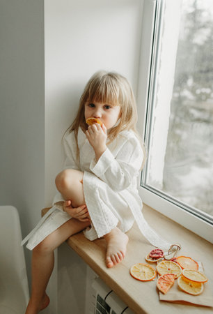 A girl in a white coat is sad sitting on the windowsill, eating fruit marshmallow.の写真素材