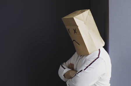 A sad man in a white shirt with a bag on his head, with a sad smiley face drawn, stands against the wall and cries.の写真素材