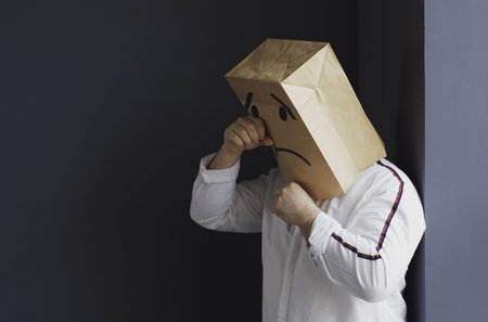 A sad man in a white shirt with a bag on his head, with a drawn crying emoticon, stands against the wall and cries. Emotions and gestures.の写真素材