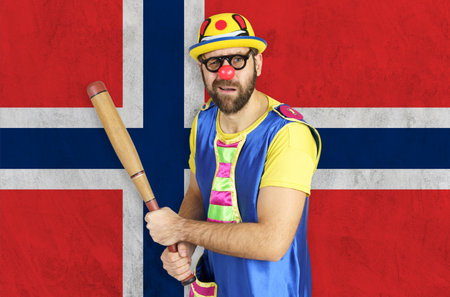 An insecure clown holds a bat in his hands against the background of the flag of Norway.の写真素材