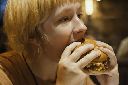 A teenager girl eats a hamburger in a restaurant, enjoying a delicious juicy burger. Close-up.の写真素材
