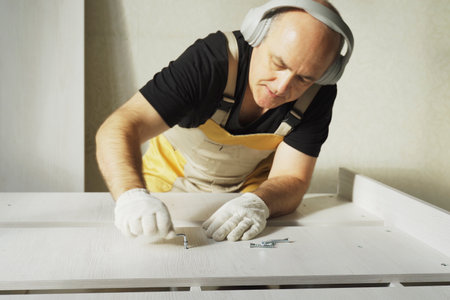 Furniture assembly. A worker fastens the parts of a bed using a confirmat. While working, he listens to music on headphones. Close-up.の写真素材
