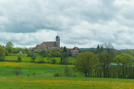 A peaceful European village sits surrounded by lush green fields punctuated by trees, with a prominent church tower rising above the clustered buildings, all under a dramatic, cloudy sky that hints at the potential for rain.の写真素材