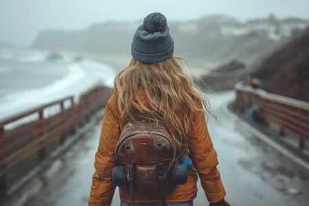 A person bundled up in a warm orange jacket and beanie walks along a foggy seaside pathway, with turbulent ocean waves in the distance.の素材