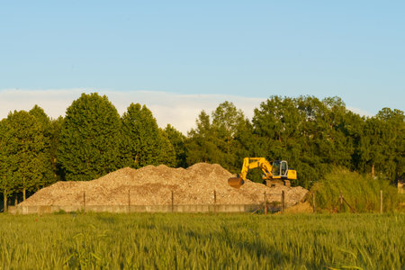 A yellow excavator stands on a pile of yellow sand on a field, with green trees in the background.