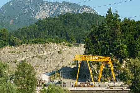 A yellow gantry crane is actively lifting materials at a quarry with a backdrop of green mountains...