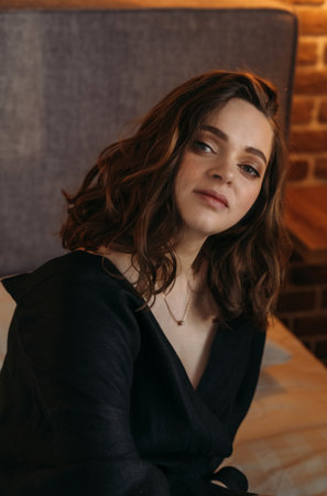 A serene young woman in a black dress sits in a cozy indoor setting, with soft ambient lighting highlighting her features. Vertical frame.の写真素材