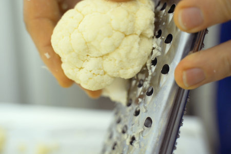 A person is grating cauliflower using a hand grater, producing finely shredded pieces. The grater is held firmly while the cauliflower is rubbed against the sharp surface.の写真素材