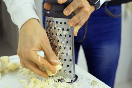 A person is grating cauliflower using a hand grater, producing finely shredded pieces. The grater is held firmly while the cauliflower is rubbed against the sharp surface.の写真素材