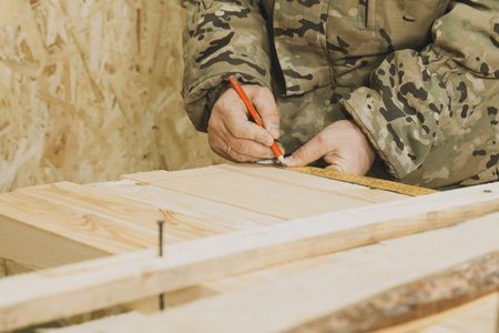 A carpenter in camouflage attire uses a tape measure and pencil to mark precise measurements on wooden planks at a workbench.の写真素材
