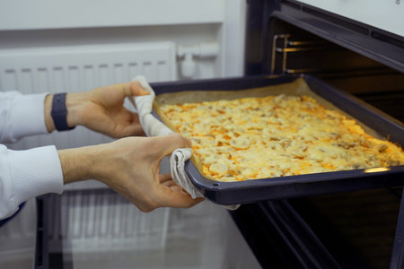 A man wearing oven mitts pulls a tray of freshly baked vegetarian pizza out of a hot oven as steam...