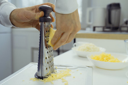 A person is grating cheese using a metal grater. The grater is creating fine shreds of cheese, with the persons hand firmly holding the grater and moving it back and forth.の写真素材