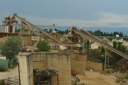 A busy mining facility with multiple conveyor belts transporting materials, large machinery, and vehicles like a cement mixer against a backdrop of a cloudy sky.の写真素材