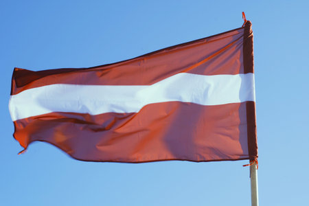 The national flag of Latvia flutters in the breeze, showing its distinctive carmine and white stripes under a cloudless sky.の写真素材