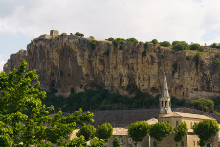 A church with a tall spire sits among buildings at the base of a large limestone cliff in a rural French town.の写真素材