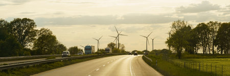 A paved road winds through a rural landscape with trees lining the sides and a line of wind turbines in the distance against a clear, sunset sky.の写真素材