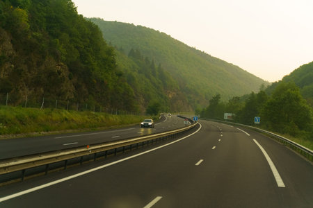A scenic mountain road curves through lush greenery on a misty morning. Cars pass by, creating a...
