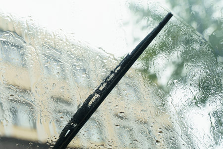 Raindrops cascade down a car windshield, distorting the cityscape beyond. The windshield wipers move rhythmically, fighting the downpour during a tranquil afternoon.の写真素材