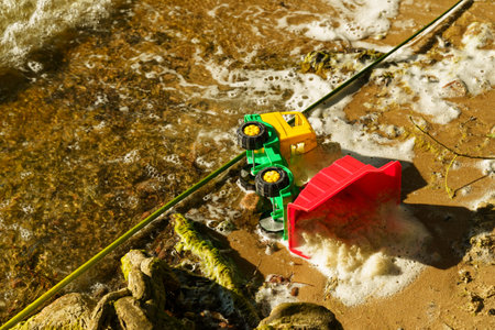 A bright yellow and green toy truck rests on a sandy beach, partially submerged in foamy water,...