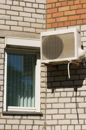 An air conditioning unit is securely fastened to a brick wall, situated next to a window with white blinds, showing a bright and warm day outside, reflecting modern urban living.の写真素材