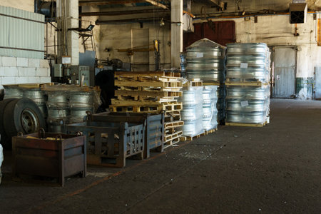 Inside an expansive industrial warehouse, piles of metallic barrels and wooden pallets are neatly organized, showcasing a busy atmosphere of storage and logistics during daylight.の写真素材