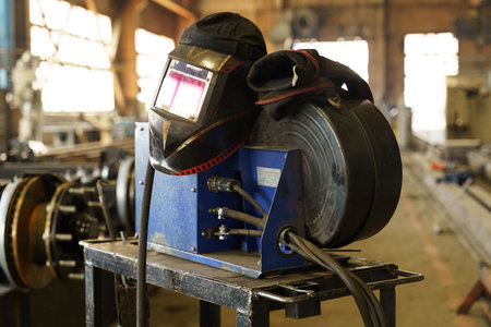A welding helmet rests atop a blue welding machine on a sturdy table in an industrial workshop....