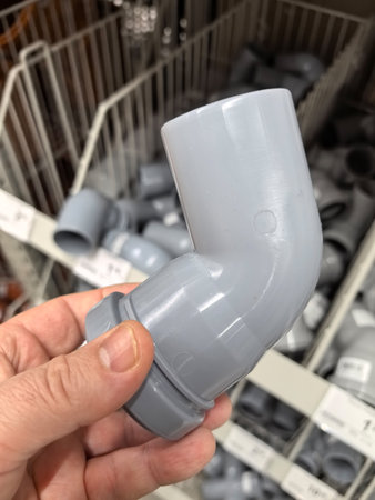 A person holds a gray plumbing elbow fitting in a hardware store aisle filled with various supplies. Bright lighting highlights the organization of the items on display, showing DIY potential.の写真素材