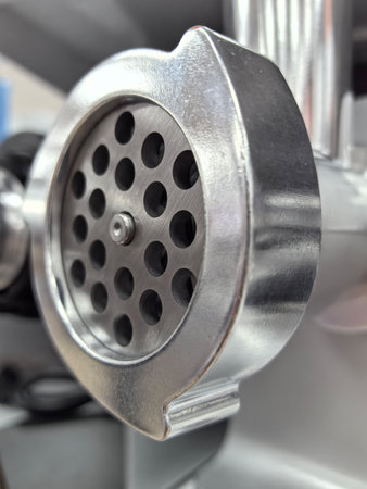 A close-up of a metal meat grinder's plate showcases its round holes and textured design. The shiny surface reflects light as it stands out against a modern kitchen background.の写真素材