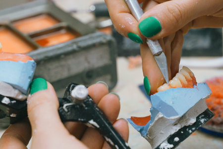 A dental technician is focused on creating a denture, precision cutting the mold while processing a wax model. Tools and materials are meticulously arranged in a well-equipped workshop setting.の写真素材