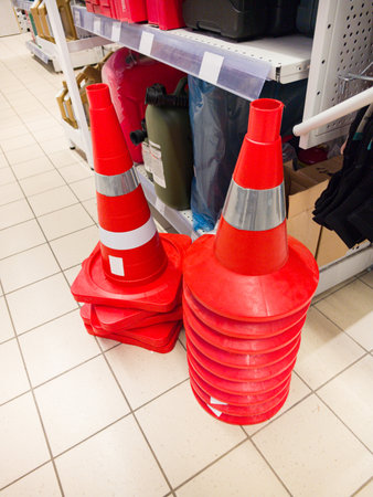 A vibrant display of orange traffic cones rests against a shelving unit in a bustling retail environment. Shoppers pass by, unaware of the colorful safety tools waiting for use.の写真素材