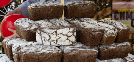 Stacks of rustic dark bread with a cracked surface sit at a market stall. Nearby, people gather, enjoying the vibrant atmosphere of the autumn celebration, engaging in local traditions.の写真素材