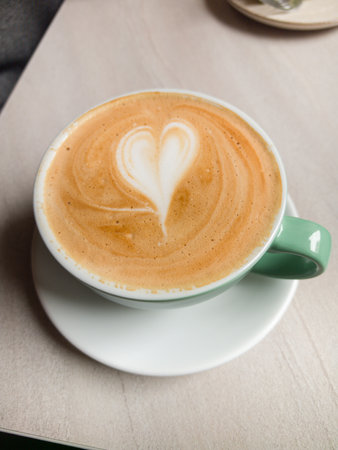 A beautifully crafted cup of coffee featuring a heart design sits on a white saucer in a charming cafe. Natural light enhances the warm tones, creating an inviting atmosphere for relaxation.の写真素材