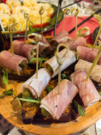 A wooden platter displays an assortment of rolled meats, garnished with herbs and served on a slice of bread. The vibrant colors of nearby dishes create a joyful atmosphere.の写真素材