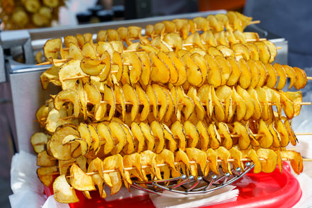Golden spiral-cut potatoes are skewered and perfectly fried, enticing visitors at a bustling outdoor food market during a sunny afternoon. The aroma fills the air, drawing food lovers.の写真素材